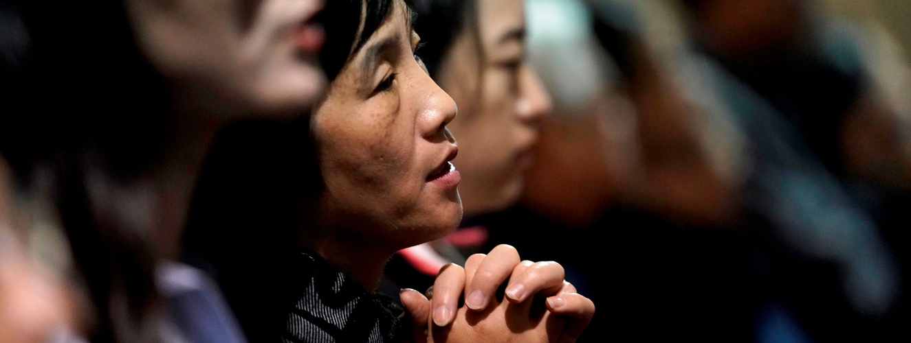 Eine Frau sitzt in einer vollen Kirchenbank mit gefalteten Händen und betet andächtig