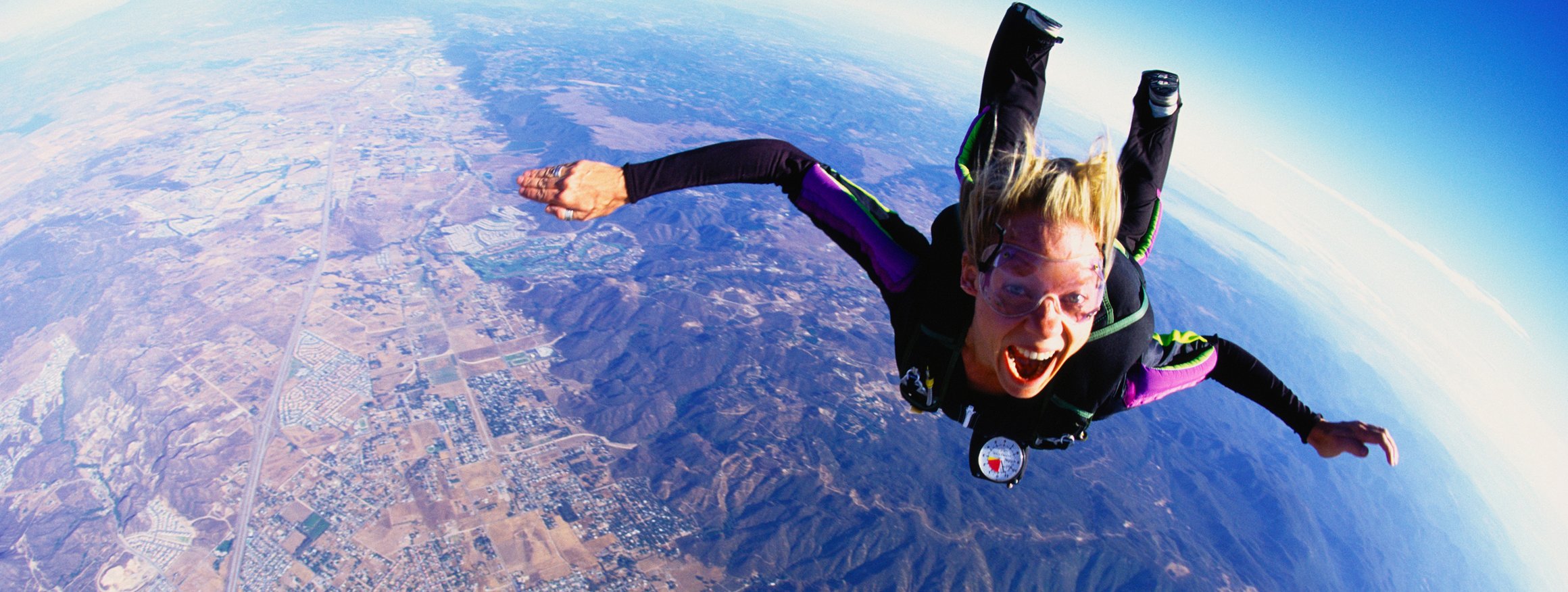 Eine Fallschirmspringerin ist in der Luft im freien Fall mit ausgestreckten Armen und Beinen und lacht dabei.