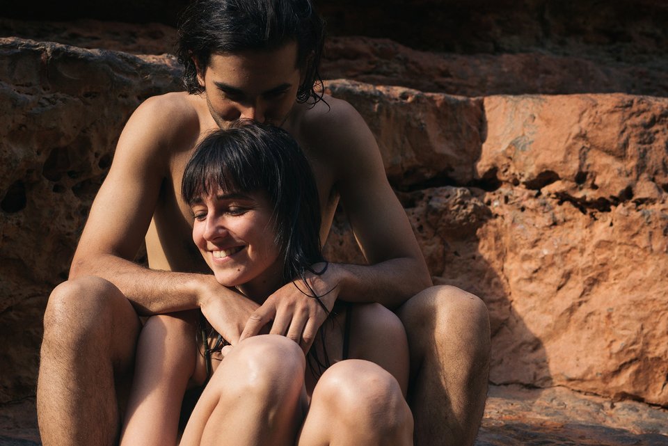 Ein junges nacktes Paar sitzt lachend vor einer Felswand, während er die Arme um ihre Schultern gelegt hat.