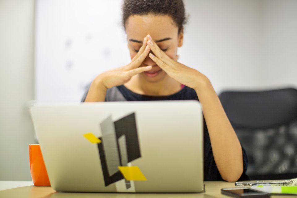 Eine junge Frau sitzt vor ihrem Laptop, die Hände vor das Gesicht gefaltet und wirkt müde