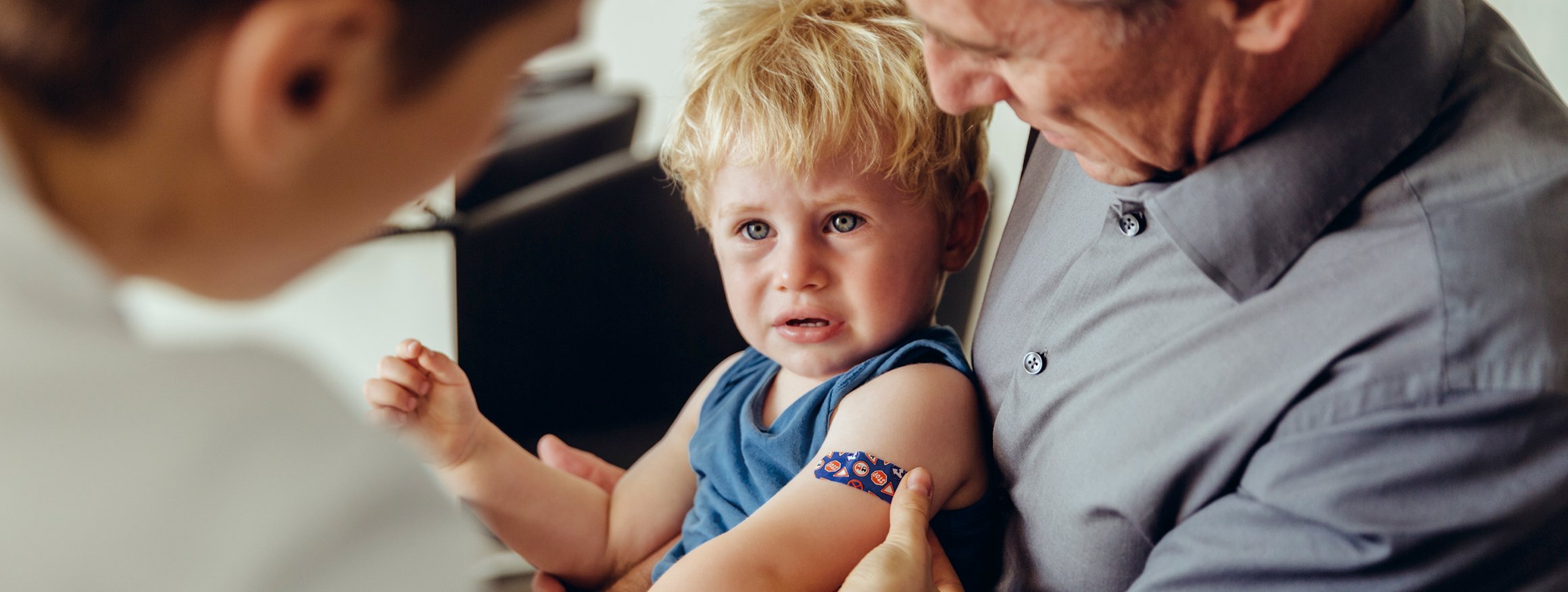 Ein Kind sitzt weinend auf dem Schoss des Vaters, während der Arzt in weißem Kittel dem Kind ein buntes Pflaster nach einer Impfung auf den Arm klebt
