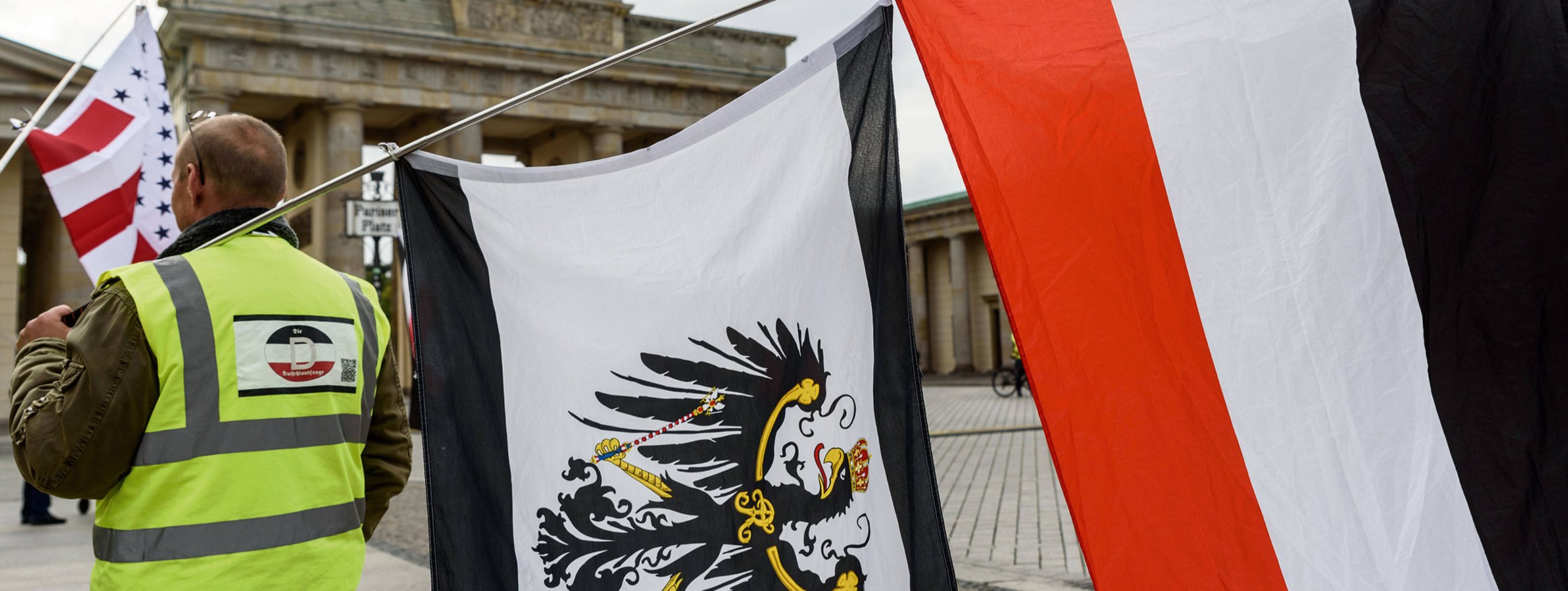 Ein Reichsbürger trägt auf einer Demonstration am Brandenburger Tor eine gelbe Weste mit Deutschland-Symbol und zwei Flaggen