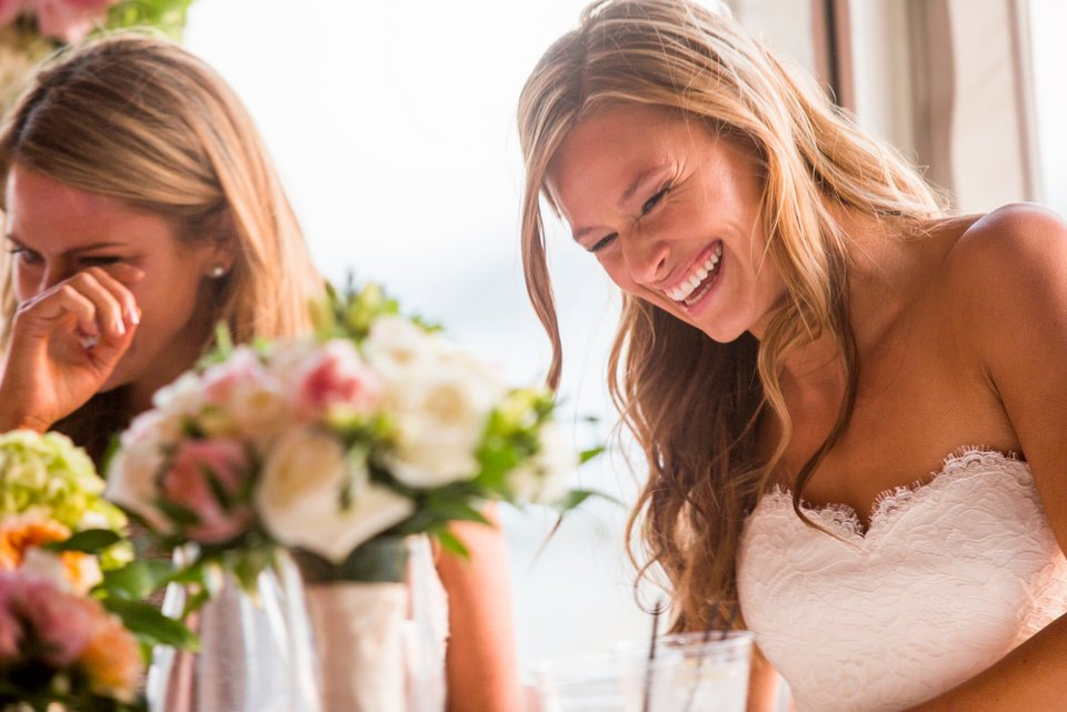 Braut im Brautkleid an einem Tisch mit Blumengestecken, lacht glücklich und ein weiblicher Hochzeitsgast wischt sich vor Rührung eine Träne aus dem Auge