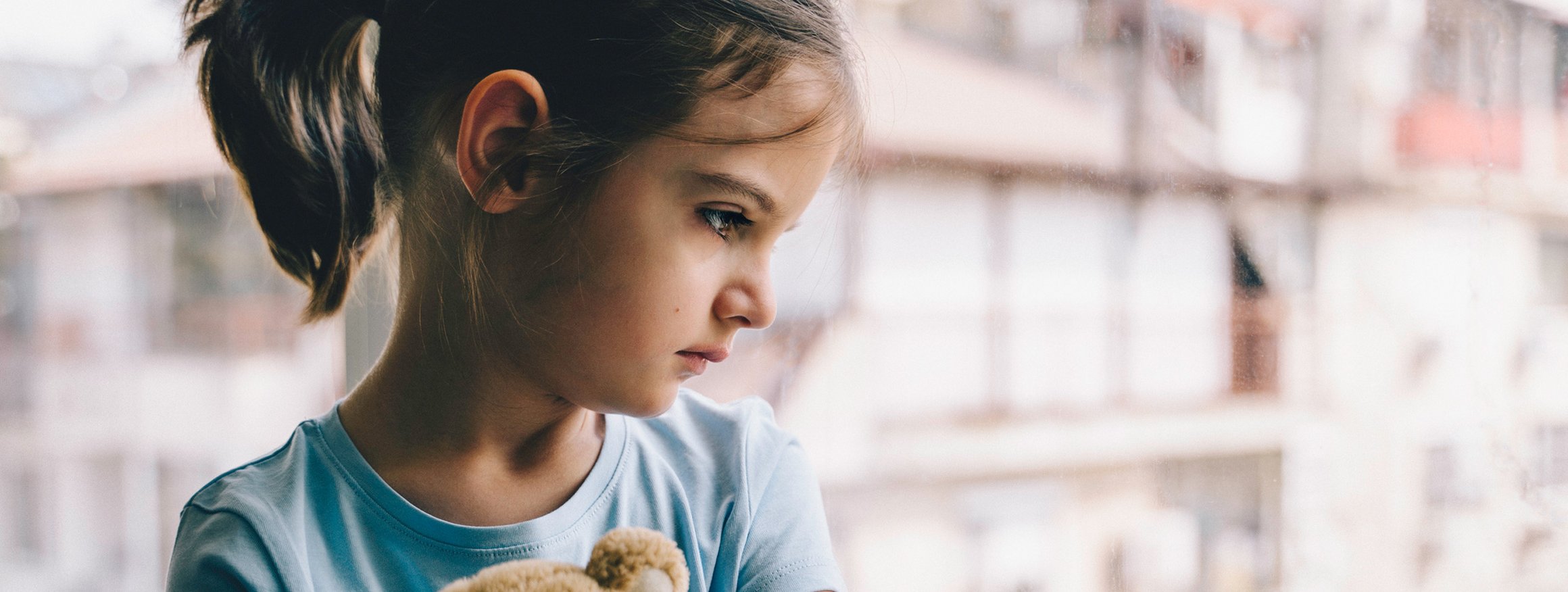 Ein kleines Mädchen hält ihren Teddy im Arm und schaut traurig aus dem Fenster