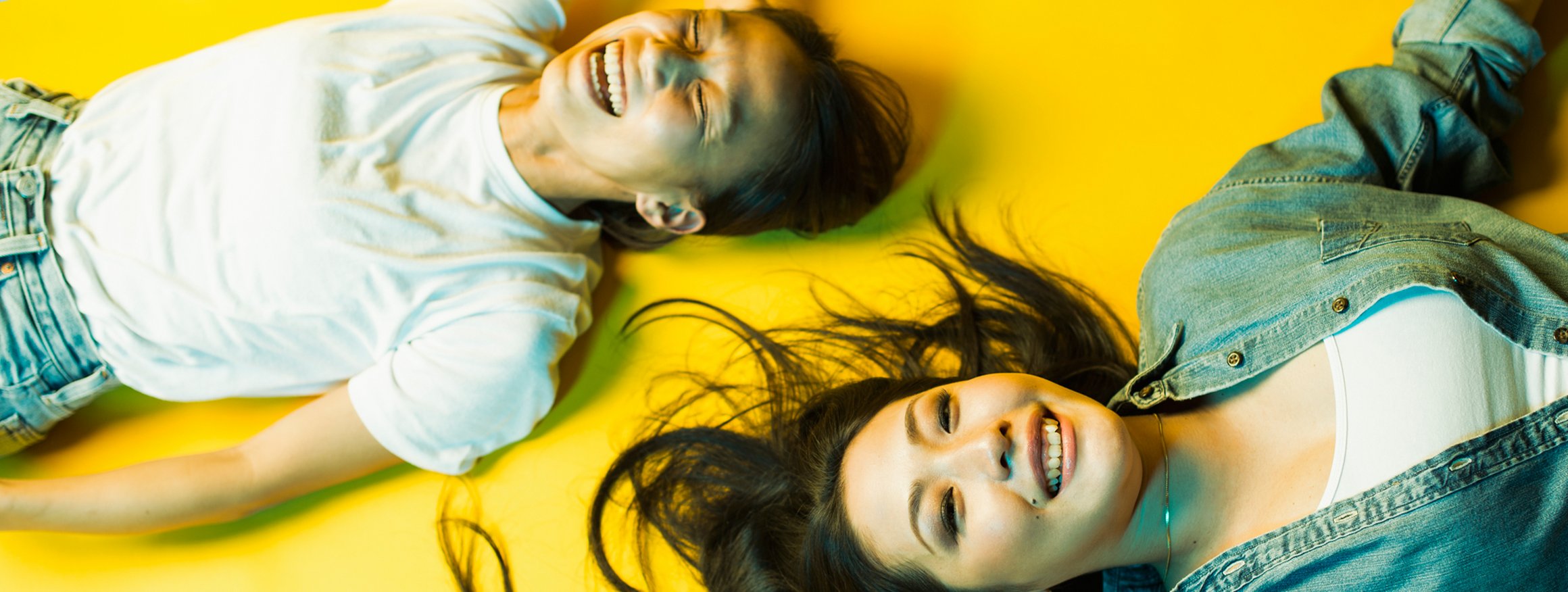 Zwei junge Frauen aus Japan liegen auf einem gelben Boden und lachen ausgelassen