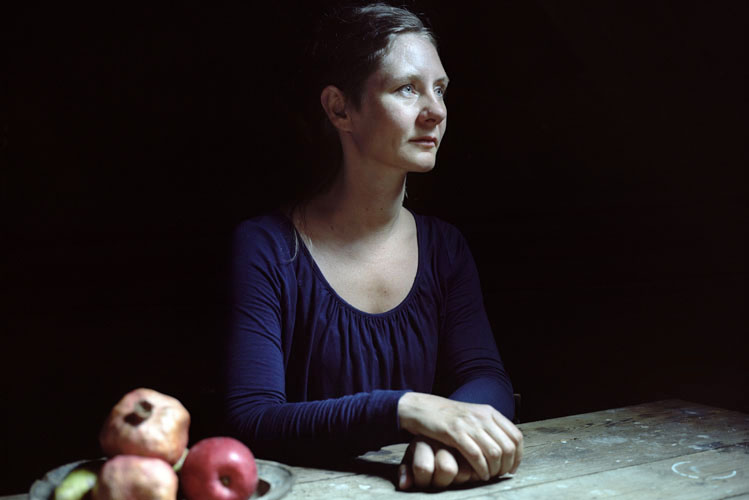 Eine Frau sitzt depressiv und alleine an einem Holztisch mit Obstschale im Hintergrund ist Dunkelheit