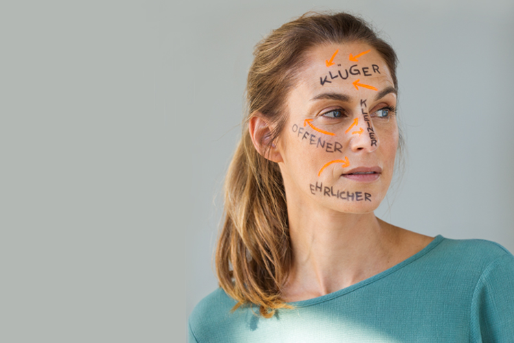Ein Frau mit gemalten Wörtern auf dem Gesicht, wie zum Beispiel "Klüger", "Offener" und "Ehrlicher" schaut voller Zweifel in die Ferne