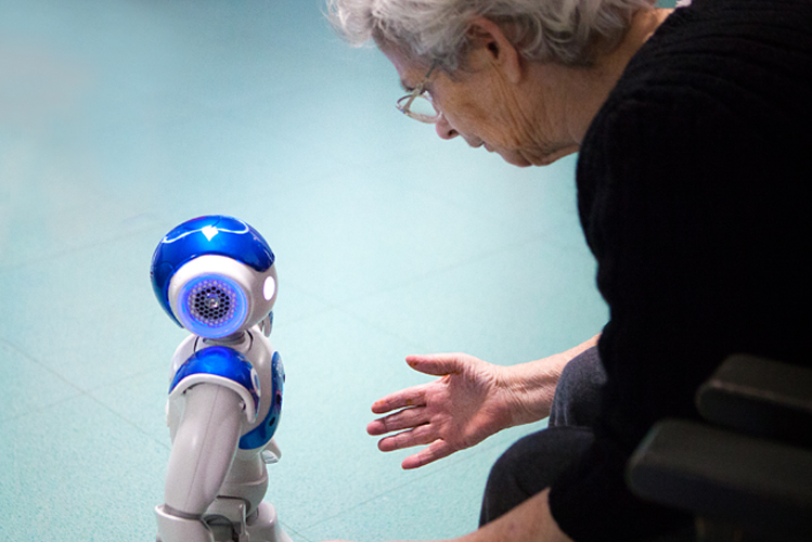 Foto zeigt eine alte Frau im Pflegeheim, die sich zu einem Roboter herunterbeugt