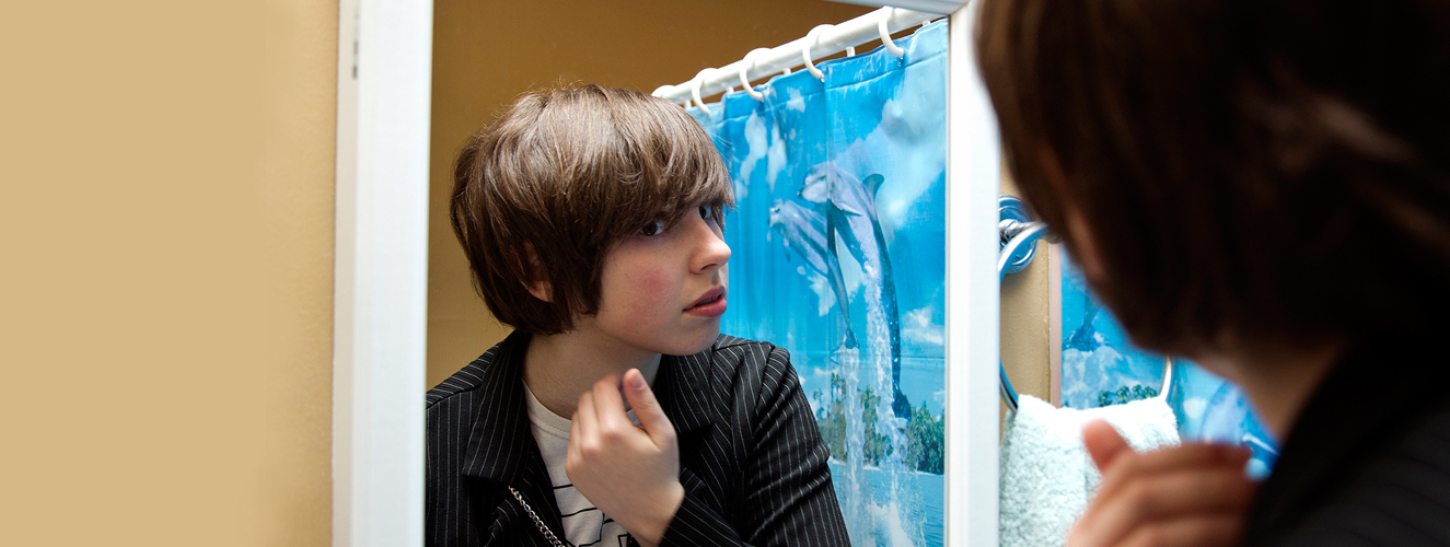 Ein androgyn aussehender Jugendlicher schaut unsicher in den Spiegel