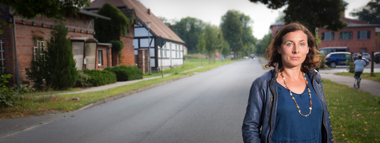 Die Schriftstellerin Juli Zeh steht in einer dörflichen Idylle an der Straße und blickt nachdenklich