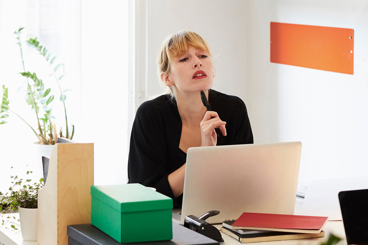 Eine junge Frau sitzt nachdenklich an ihrem Arbeitsplatz am PC und ist dabei ganz fokussiert auf die Arbeit