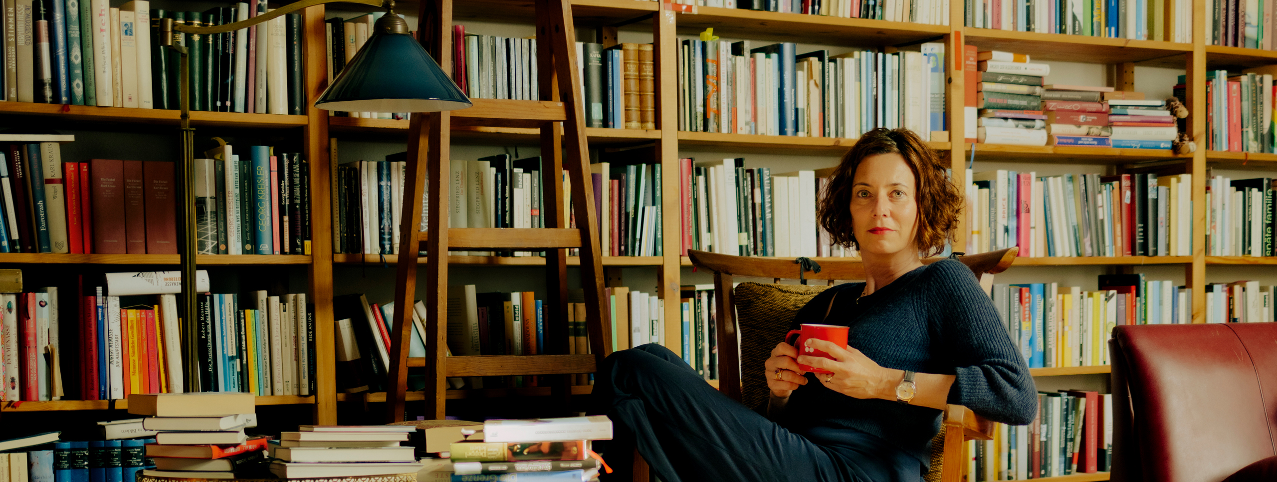 Die Schriftstellerin Eva Menasse, sitzt lässig auf einem Stuhl vor einer großen Bücherwand und hält dabei eine Tasse in der Hand