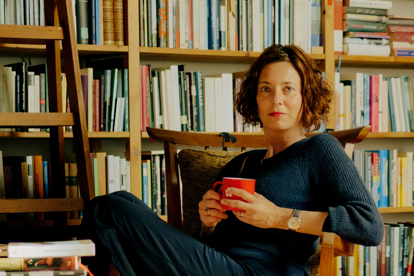 Die Schriftstellerin Eva Menasse sitzt lässig, mit einer Tasse in der Hand, vor ihrer Bücherwand