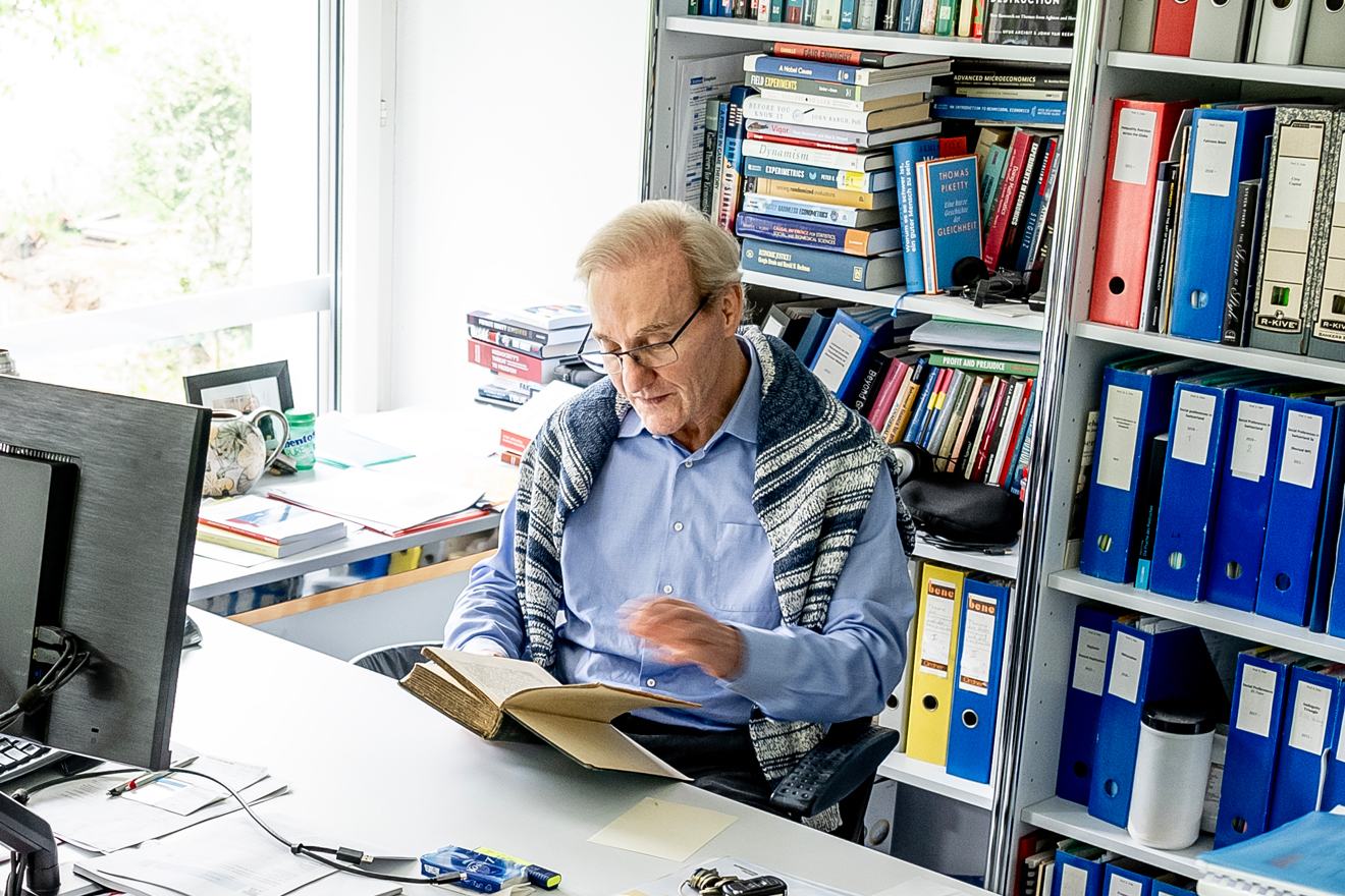 Der Wirtschaftswissenschaftler und Professor, Ernst Fehr sitzt an seinem Schreibtisch vor einer Bücher- und Aktenwand und liest in einem Buch