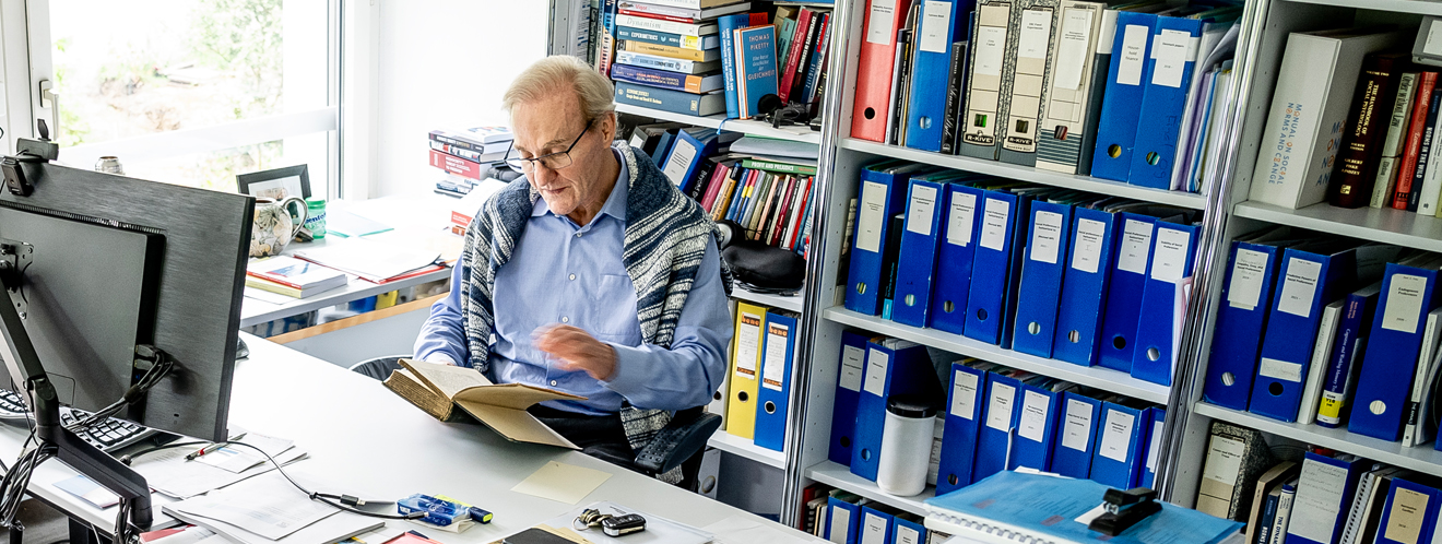Der Wirtschaftswissenschaftler und Professor, Ernst Fehr sitzt an seinem Schreibtisch vor einer Bücher- und Aktenwand und liest in einem Buch