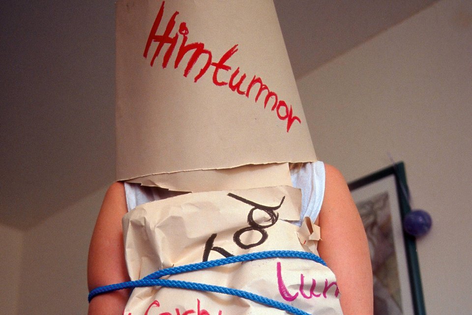 Eine Frau ist gefesselt und trägt eine Papiertüte über dem Kopf auf dem das Wort Hirntumor steht und über ihren Körper ist ebenfalls Papier mit Wörtern, wie zum Beispiel Tod