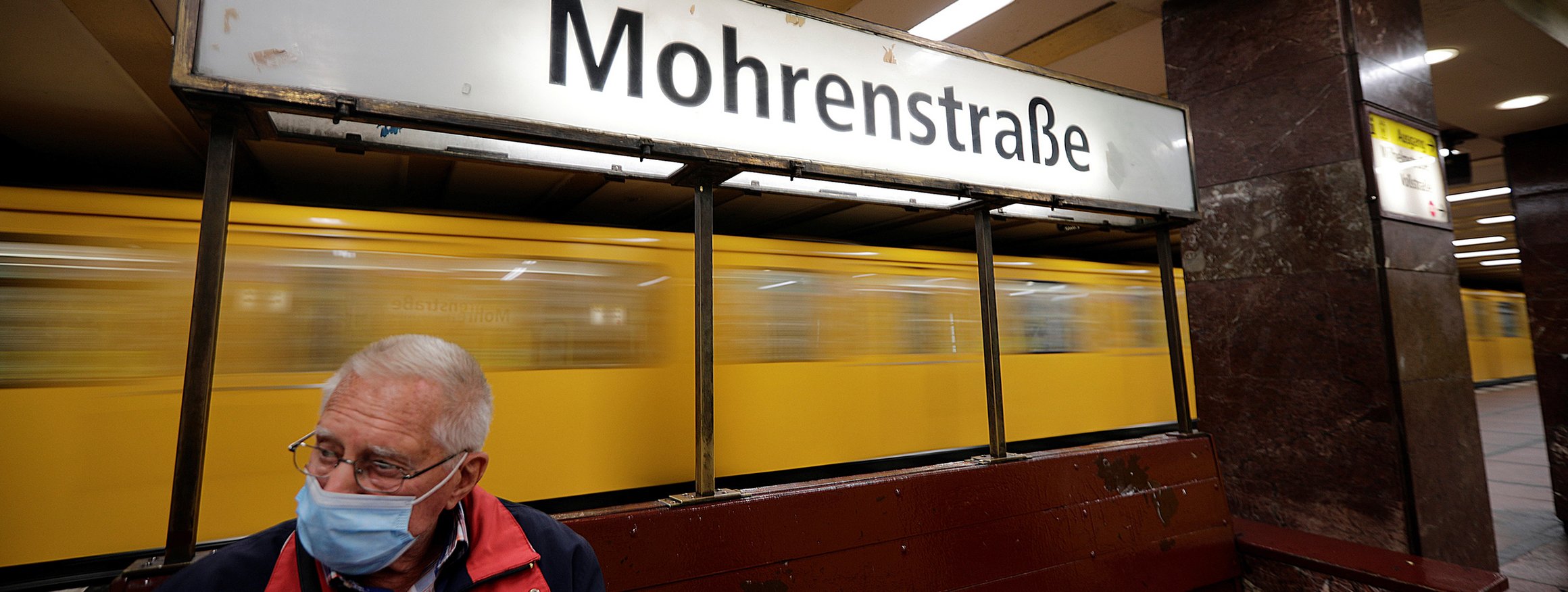 Ein älterer Mann mit Coronamaske sitzt an einer Bahnstation, die Mohrenstraße heißt, während hinter ihm eine Bahn vorbeifährt