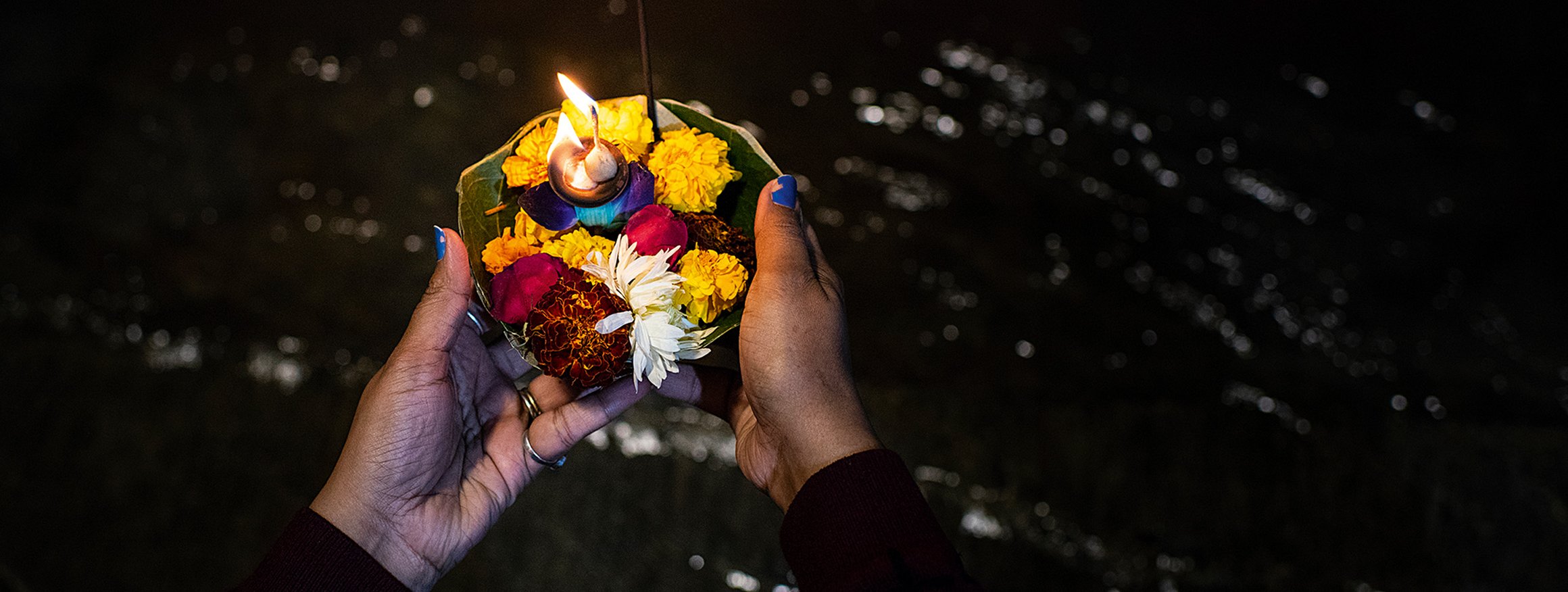 Zwei Frauenhände mit Ringen und abgeblätterten blauen Nagellack halten eine Schale mit Licht und Blumen