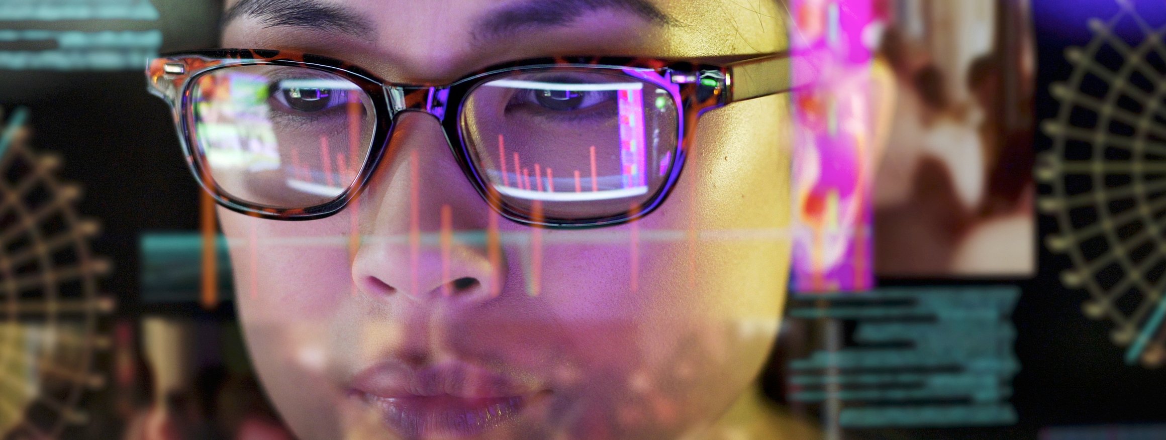 Eine junge Frau mit schwarz umrandeter Brille steht an einer Scheibe in einem Datenraum und spiegelt sich