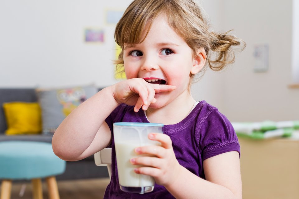 Ein kleines Mädchen hält ein Glas Milch in der Hand und lächelt verschmitzt