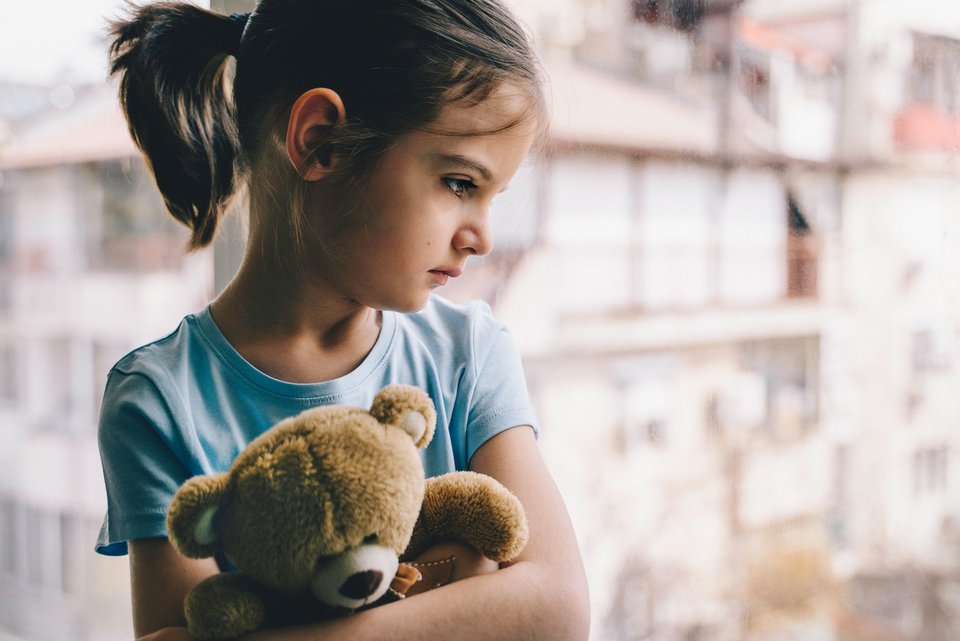 Ein kleines Mädchen hält ihren Teddy im Arm und schaut traurig aus dem Fenster