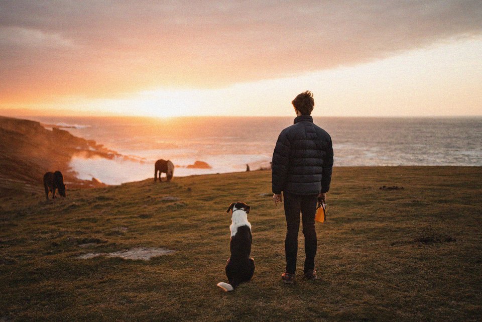 Eine androgyne Frau steht mit ihrem Hund am Meer und schaut in die Ferne