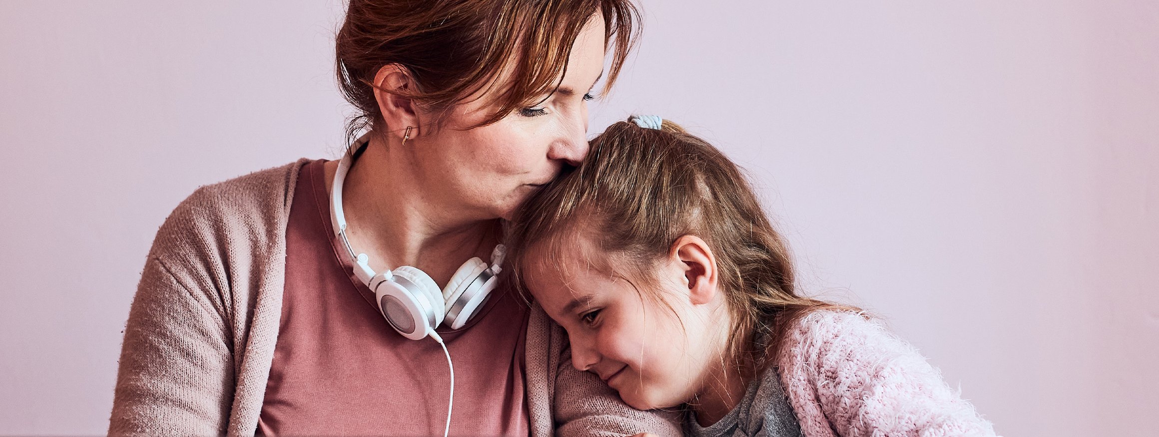 Eine alleinerziehende Mutter sitzt vor ihrem Computer im Homeoffice, daneben ihre kleine Tochter, die sich an die Mutter lehnt, die ihr fürsorglich den Kopf küsst