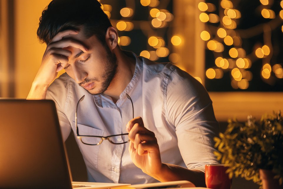 Ein junger Mann sitzt an seinem Laptop und hält sich erschöpft mit geschlossenen Augen den Kopf, während er mit einer Hand seine Brille hält