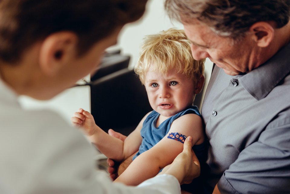 Ein Kind sitzt weinend auf dem Schoss des Vaters, während der Arzt in weißem Kittel dem Kind ein buntes Pflaster nach einer Impfung auf den Arm klebt