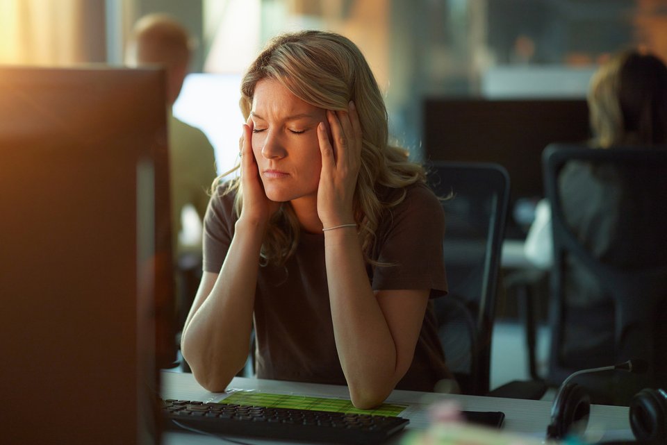 Eine Frau sitzt gestresst im Büro vor ihrem Bildschirm und hat die Augen geschlossen und die Hände an den Schläfen