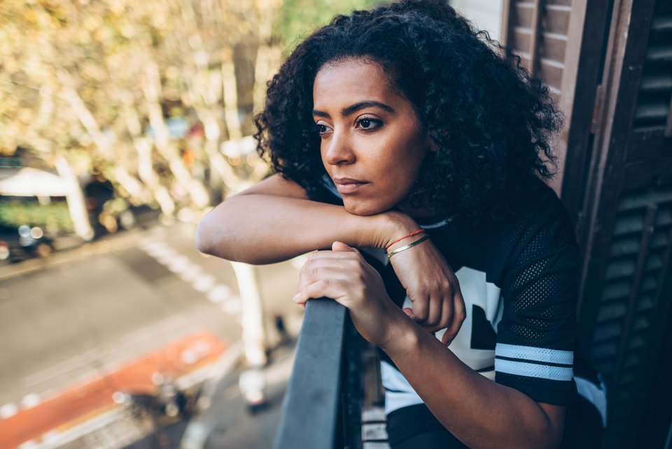 Eine junge Frau mit dunklen Locken und T-Shirt sitzt auf einem Balkon in einer Stadt und hat sich mit den Kopf traurig auf das Geländer gelegt und schaut in die Ferne