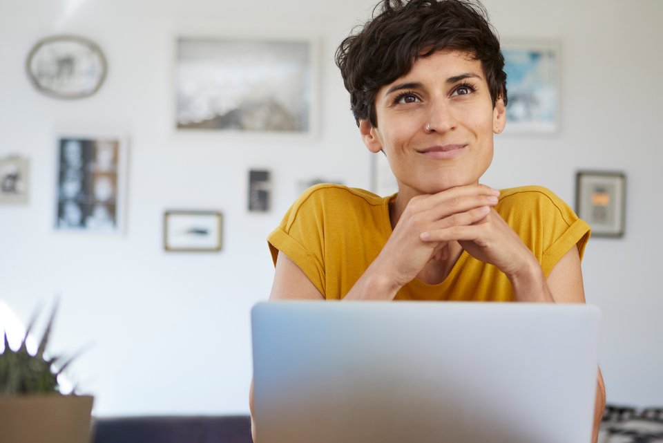 Eine junge Frau mit dunklen, kurzen Haaren und gelbem Shirt sitzt vor ihrem Laptop und denkt über sich nach