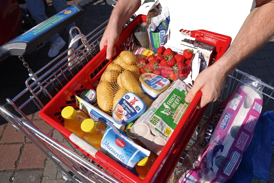 Ein Mann mit Einkaufswagen hebt seinen roten, voll gepackten, Einkaufskorb aus Plastik aus dem Wagen