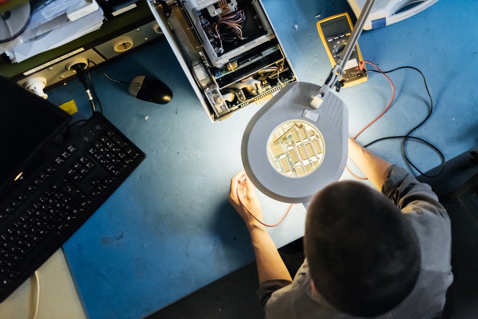 Ein Mann sitzt im Schichtbetrieb unter einer Schreibtischlampe und hat technische Geräte auf dem Tisch