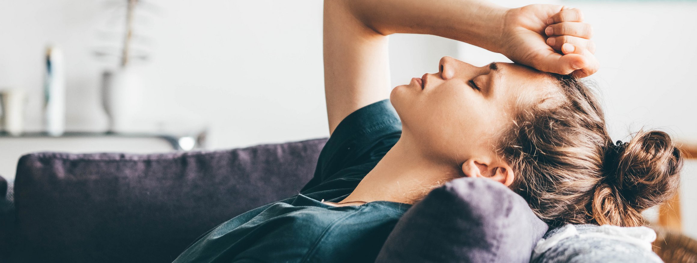 Eine junge Frau liegt auf der Couch, den Kopf auf ein Kissen gelegt, und hält sich leidend die Hand an die Stirn, während sie die Augen geschlossen hat