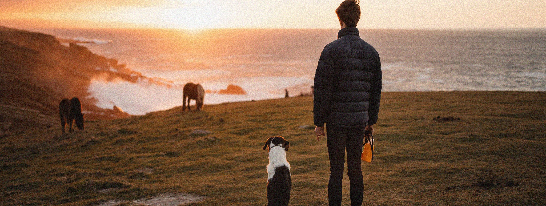 Eine androgyne Frau steht mit ihrem Hund am Meer und schaut in die Ferne