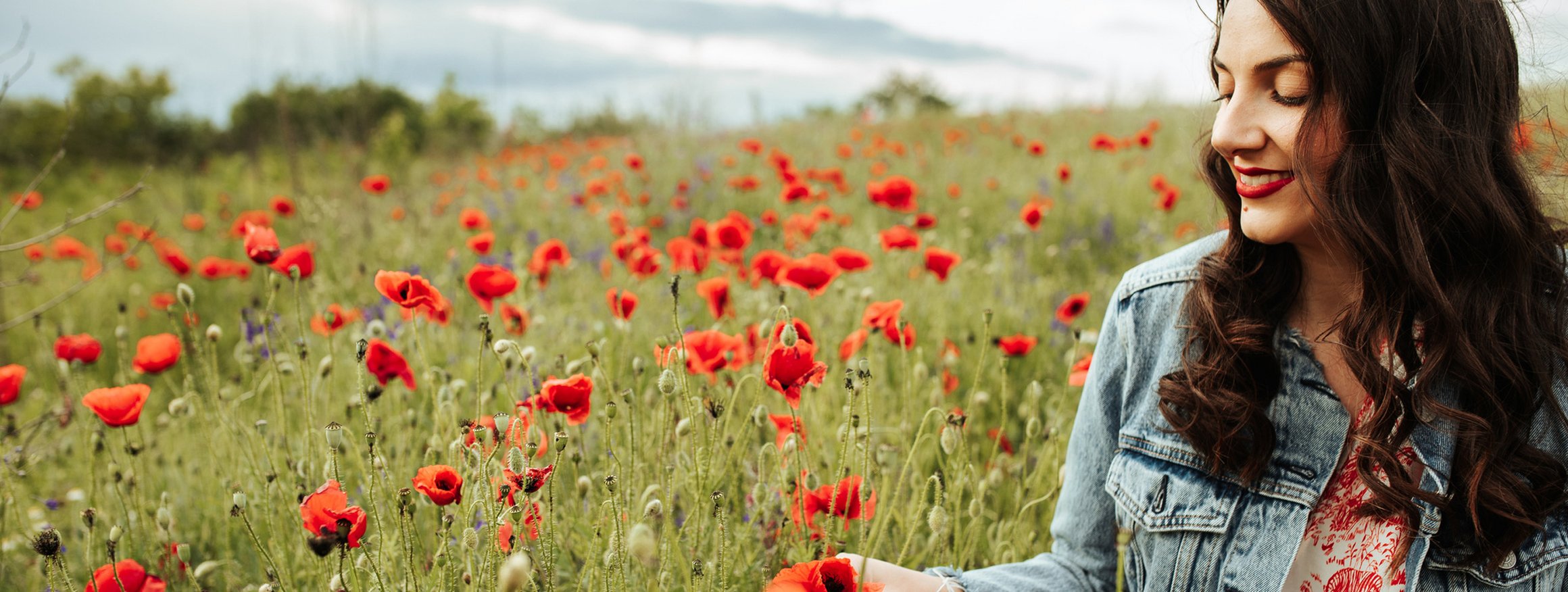 Eine junge Frau in Jeansjacke mit rotem Lippenstift und langen dunklen Haaren sitzt lächelnd in einem roten Mohnblumenfeld