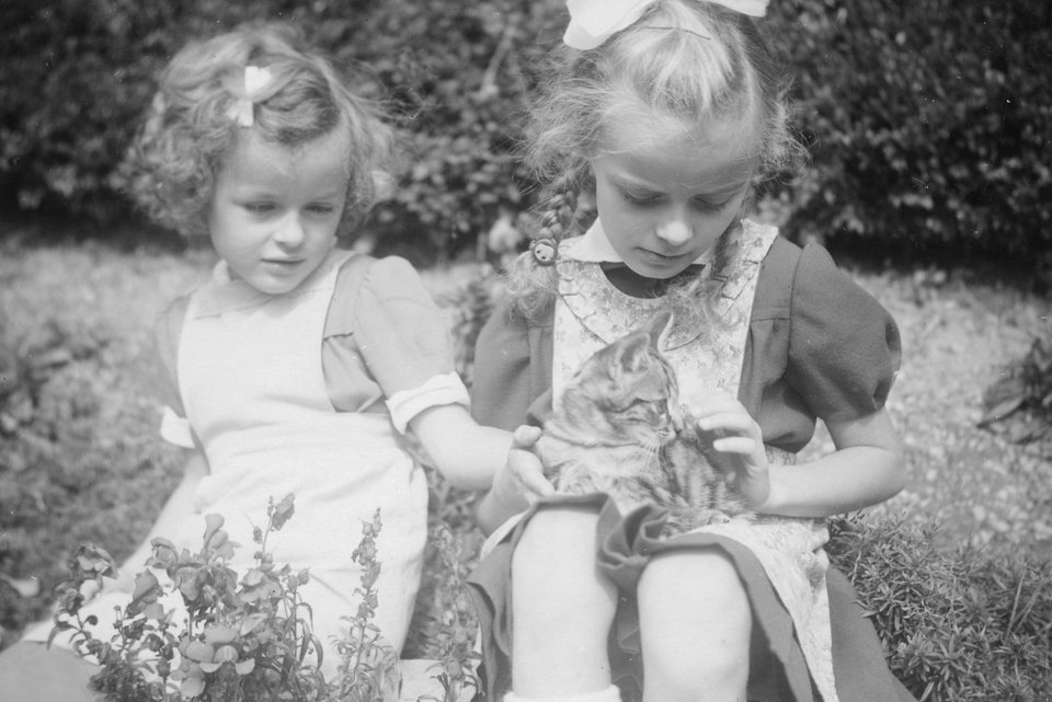 Auf einem alten schwarz-weiß Foto sind zwei blonde Mädchen mit Schürzen und Schleifen im Haar im Garten und ein Mädchen hat ein Kätzchen auf seinem Schoß
