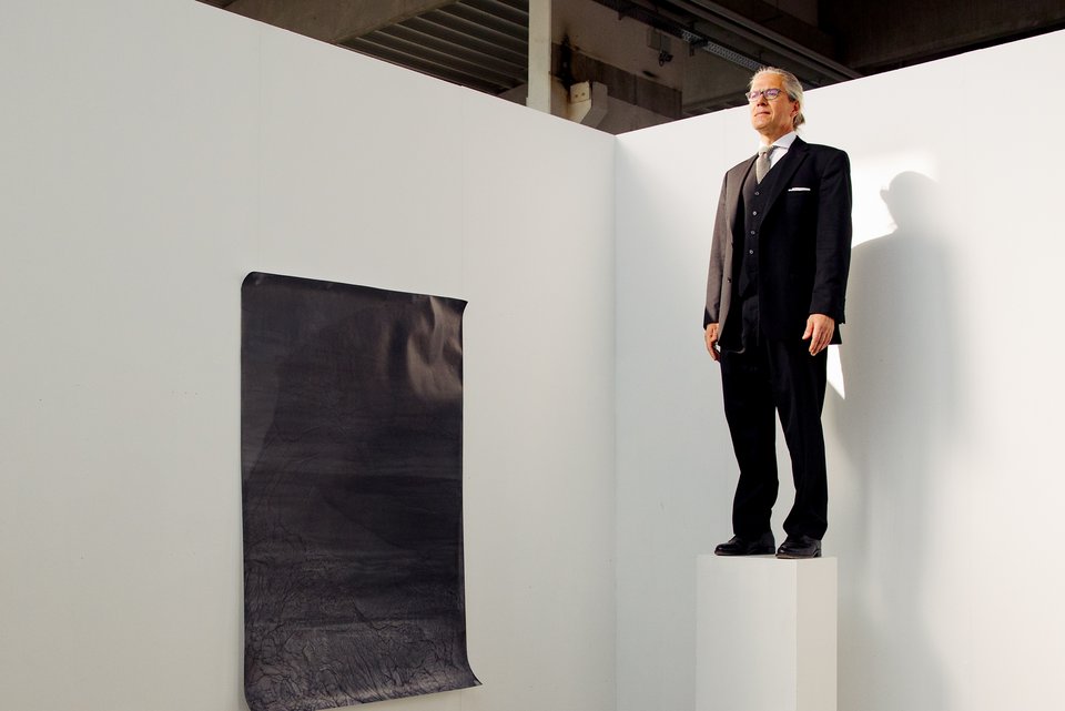 Der Schriftsteller, Steffen Kopetzky, steht in einer Kunstgalerie auf einem Podest wie eine Skulptur, daneben hängt ein Kunstwerk an der Wand