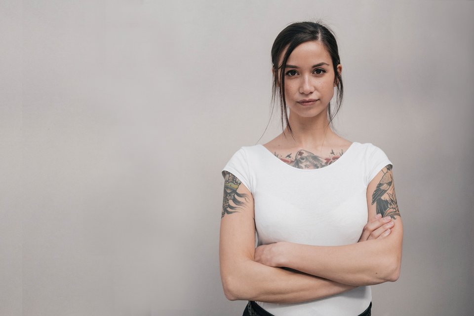 Eine junge Frau mit weißem Shirt und Tattoos schaut ernst und hat die Arme vor ihren Körper verschränkt