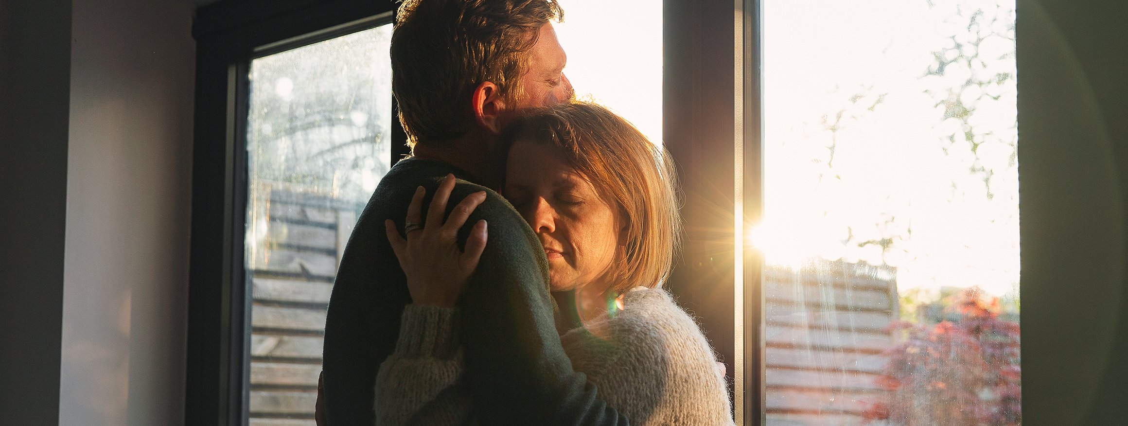 Mann und Frau umarmen sich und stehen vor einem Fenster durch das die Abendsonne scheint