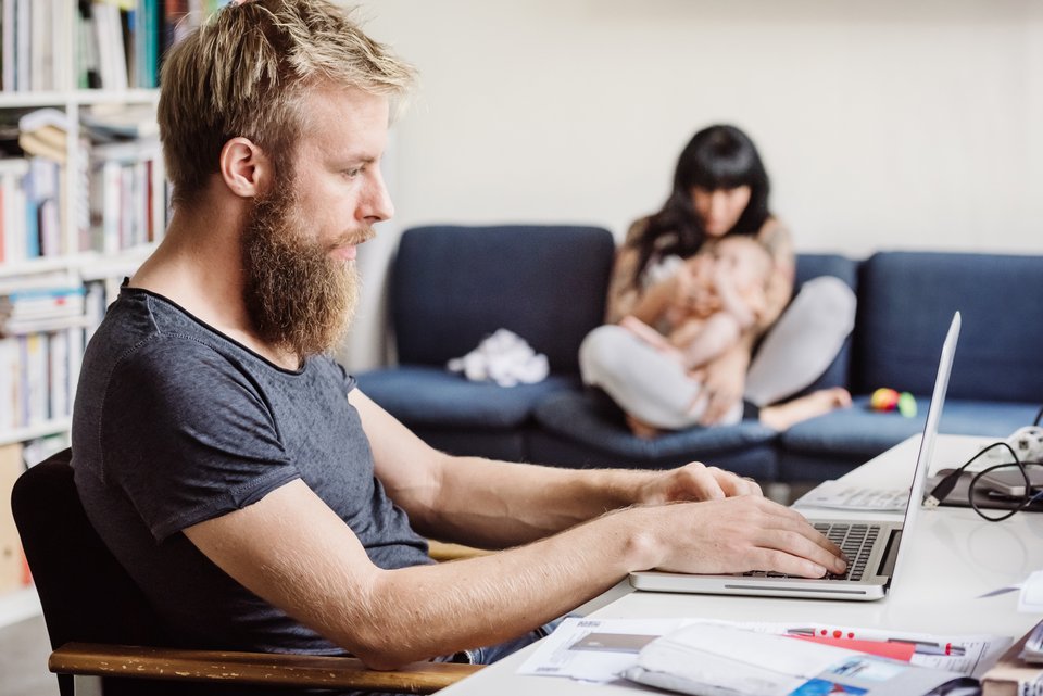 Ein jüngerer Mann sitzt in seinem Wohnzimmer im Homeoffice, dahinter sitzt seine Partnerin mit einen Baby auf dem Arm