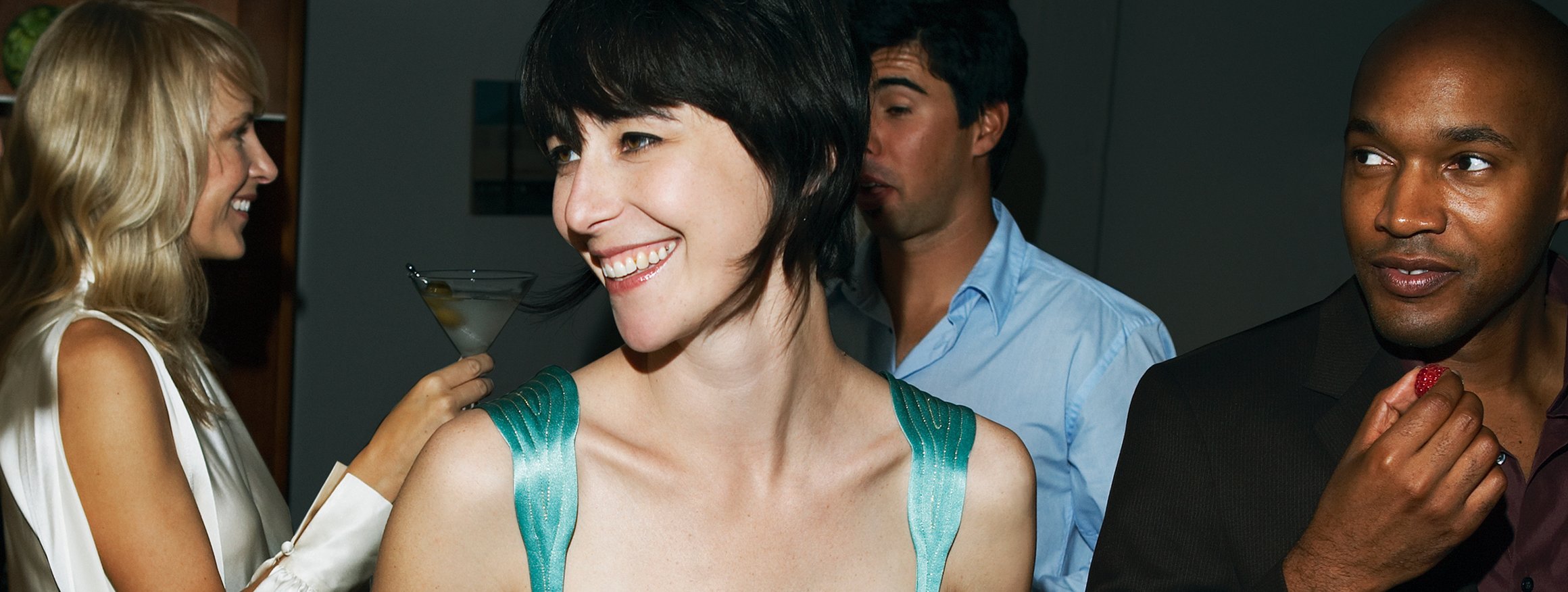Eine junge Frau steht mit einem Cocktail lachend in einer Gruppe von Menschen auf einer Party