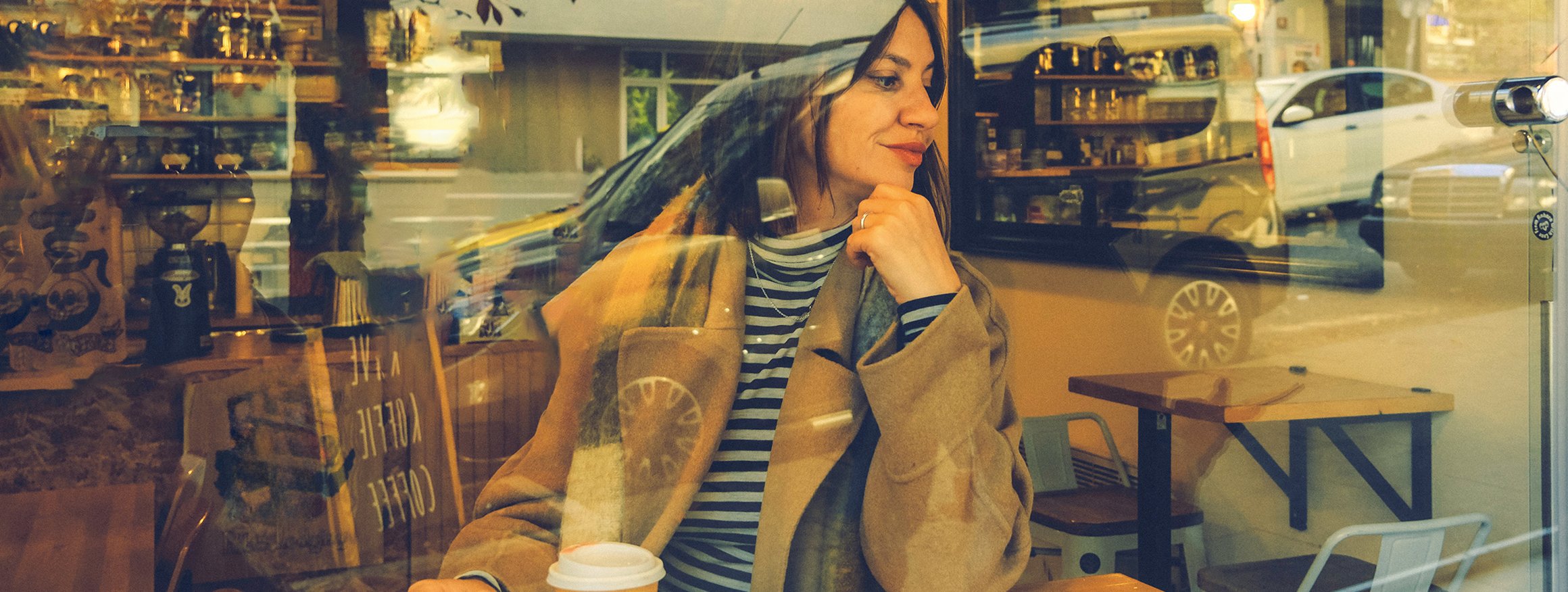 Eine junge Frau sitzt entspannt am Fenster eines Cafés, vor ihr steht ein Coffee-to-go