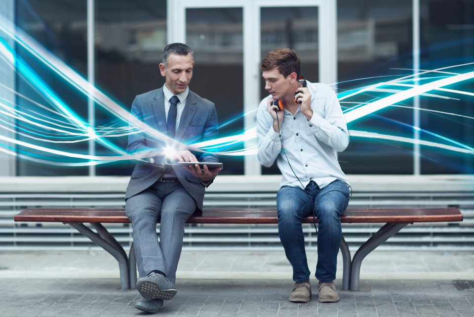 Zwei Männer sitzen vor Geschäftshaus mit Glasfenster auf einer öffentlichen Bank und schauen interessiert auf ein Tablet