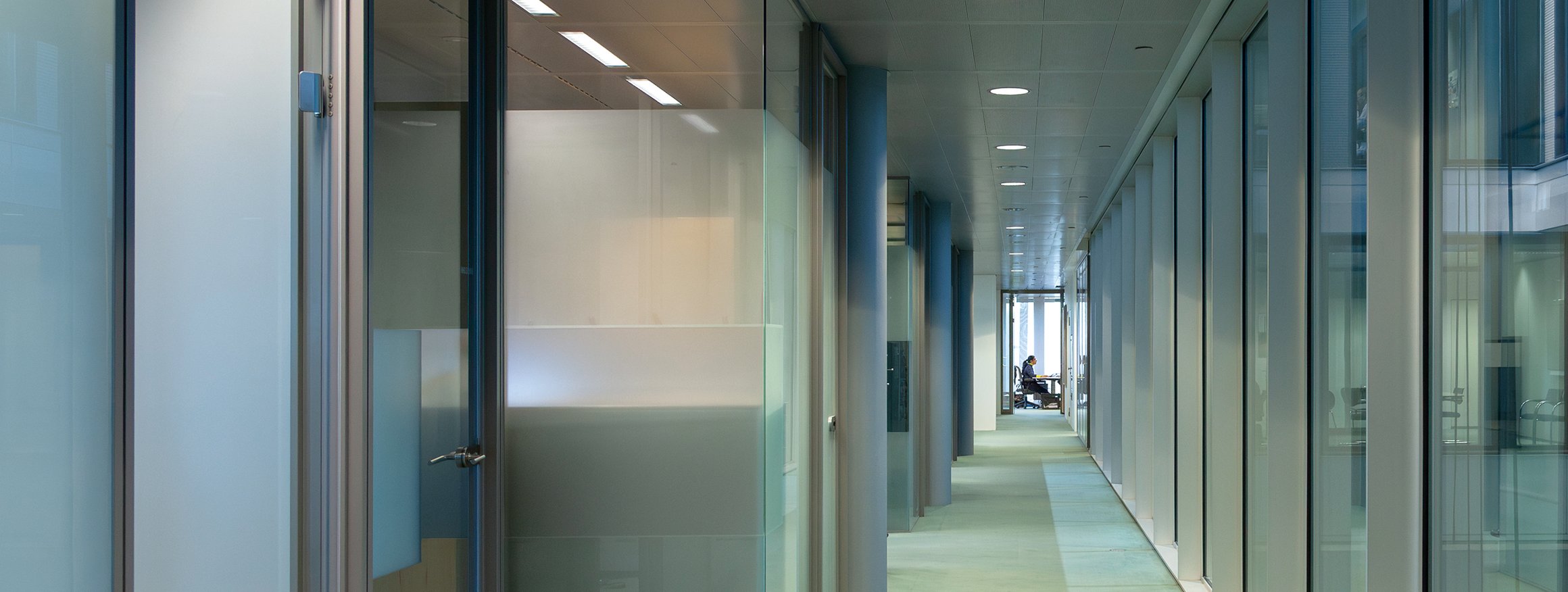 Ein sehr langer und grauer Flur, an dessen Ende ein Angestellter in einem Büro sitzt 