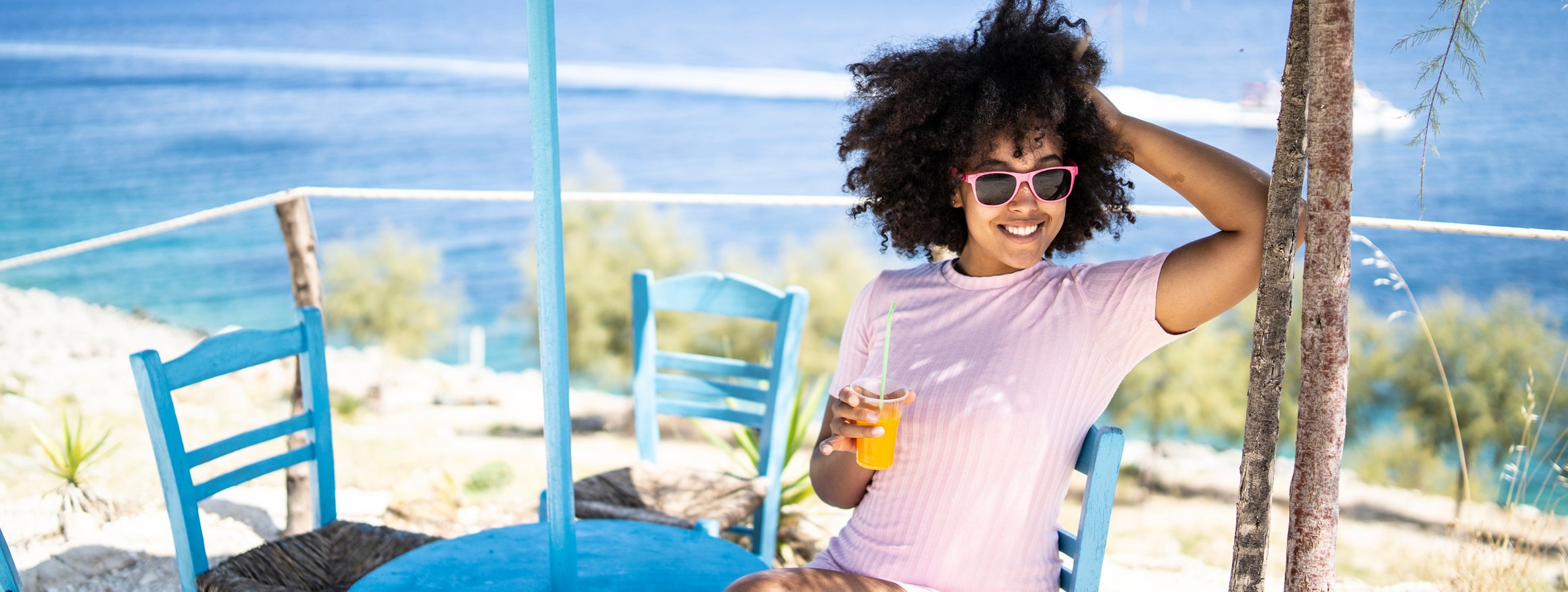 Eine junge Frau mit dunklem Lockenkopf und Sonnenbrille sitzt lachend mit einem Drink in der Hand an einer Strandbar