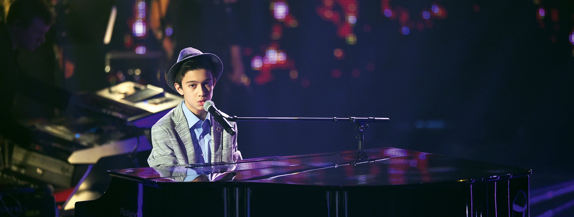 Ein Junge sitzt mit Anzug und Hut in einer Casting-Show am Klavier und singt in ein Mikro