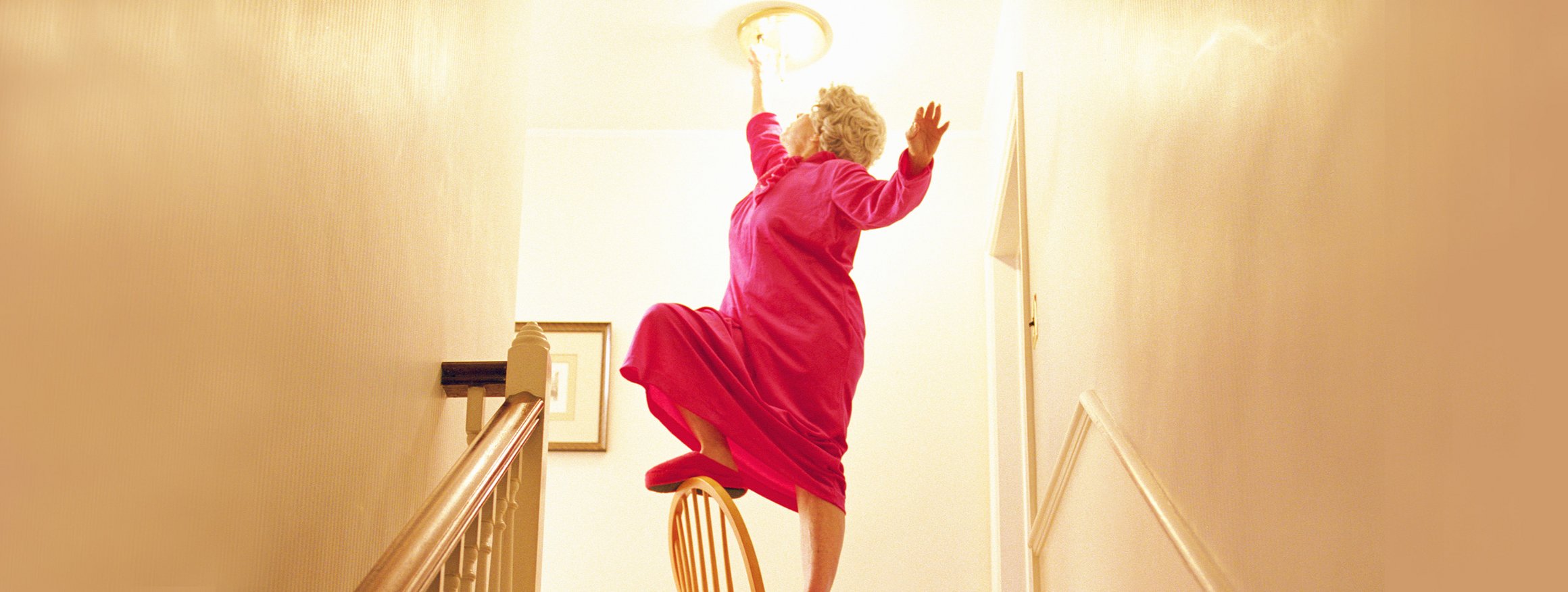 Ältere Frau balanciert auf einem Stuhl und wechselt eine Glühbirne