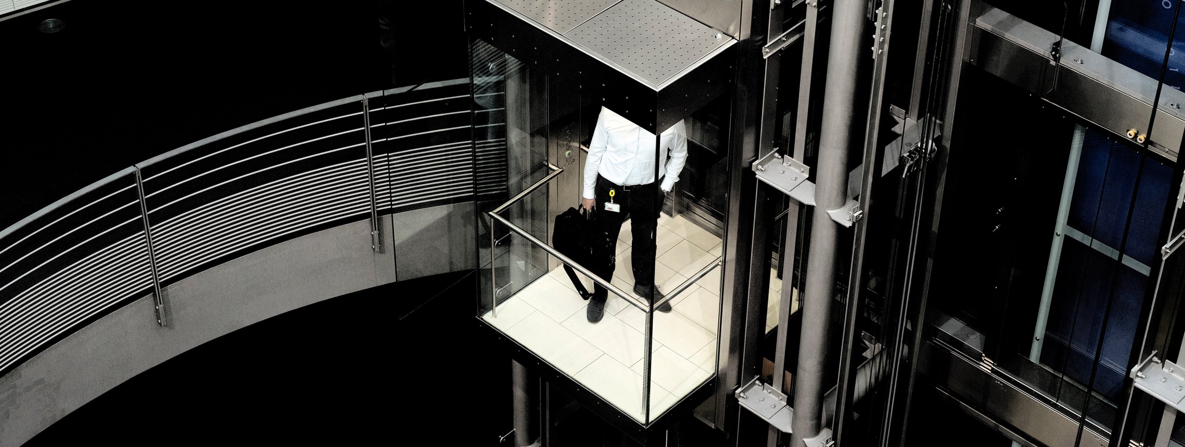 Ein Mann im Business-Look steht in einem gläsernen Aufzug in einem sehr modernen, aber kalten Bürogebäude